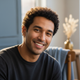 A smiling man in his late 20s, conveying trust and satisfaction. The background is blurred with dusty blue and warm cream acc